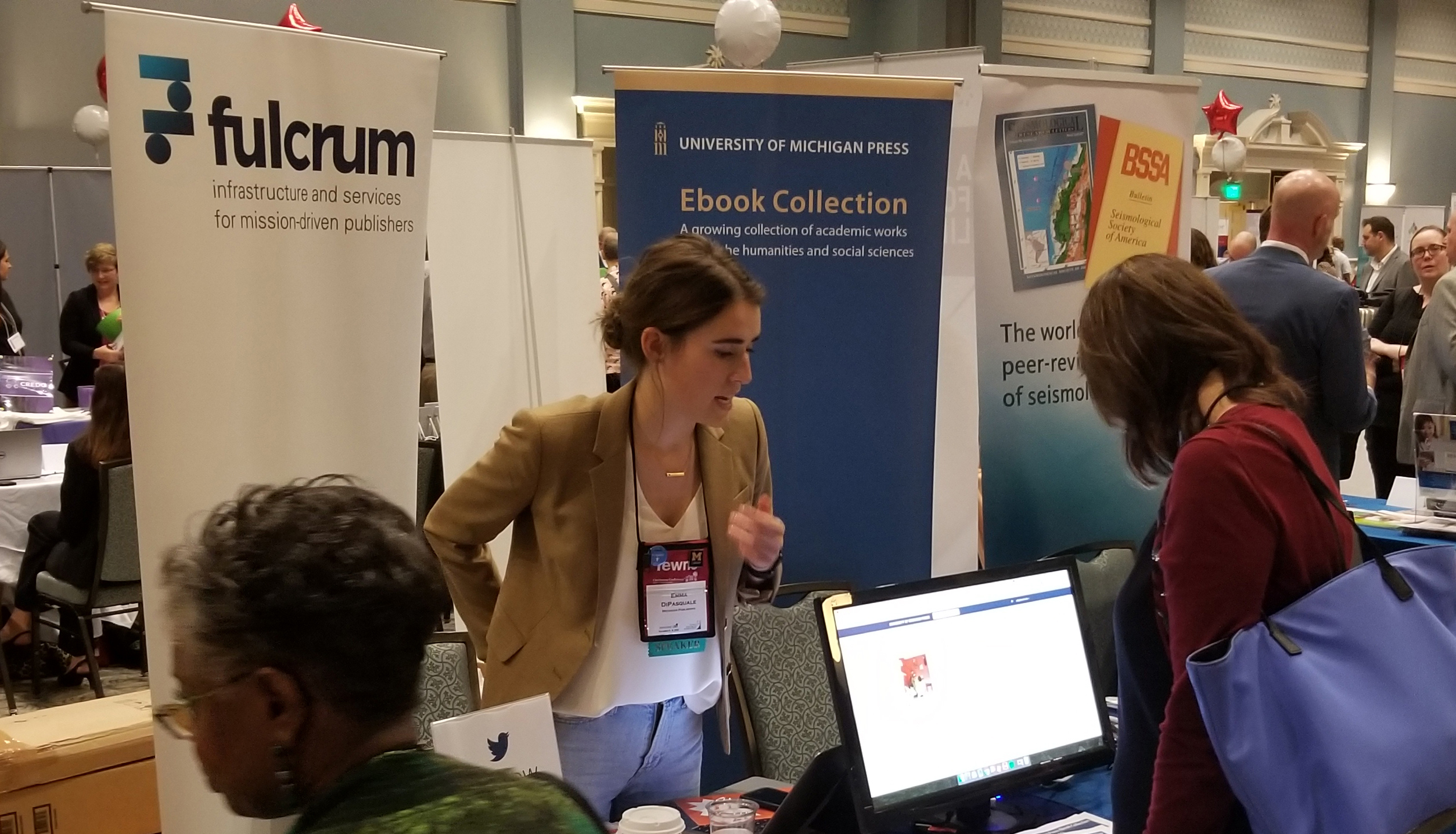 A photograph of a young woman showing a digital ebook from the University of Michigan Press Ebook collection to a Charleston conference attendee at the Michigan Publishing display booth. 
