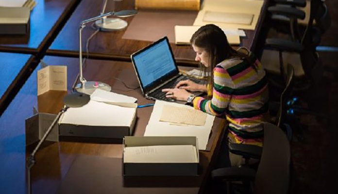 A photograph of a young woman studying with books and a computer at a table in the U-M Law Library.