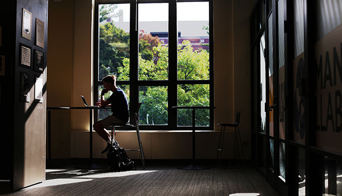 A photograph of a room covered mostly with shadow, where a student with short hair sits at a table in front of a large window with their laptop open. The tops of trees and a nearby building can be seen through the window.